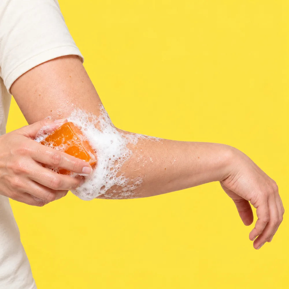 A hand rubbing the orange Asshine bar over an elbow, building a rich lather